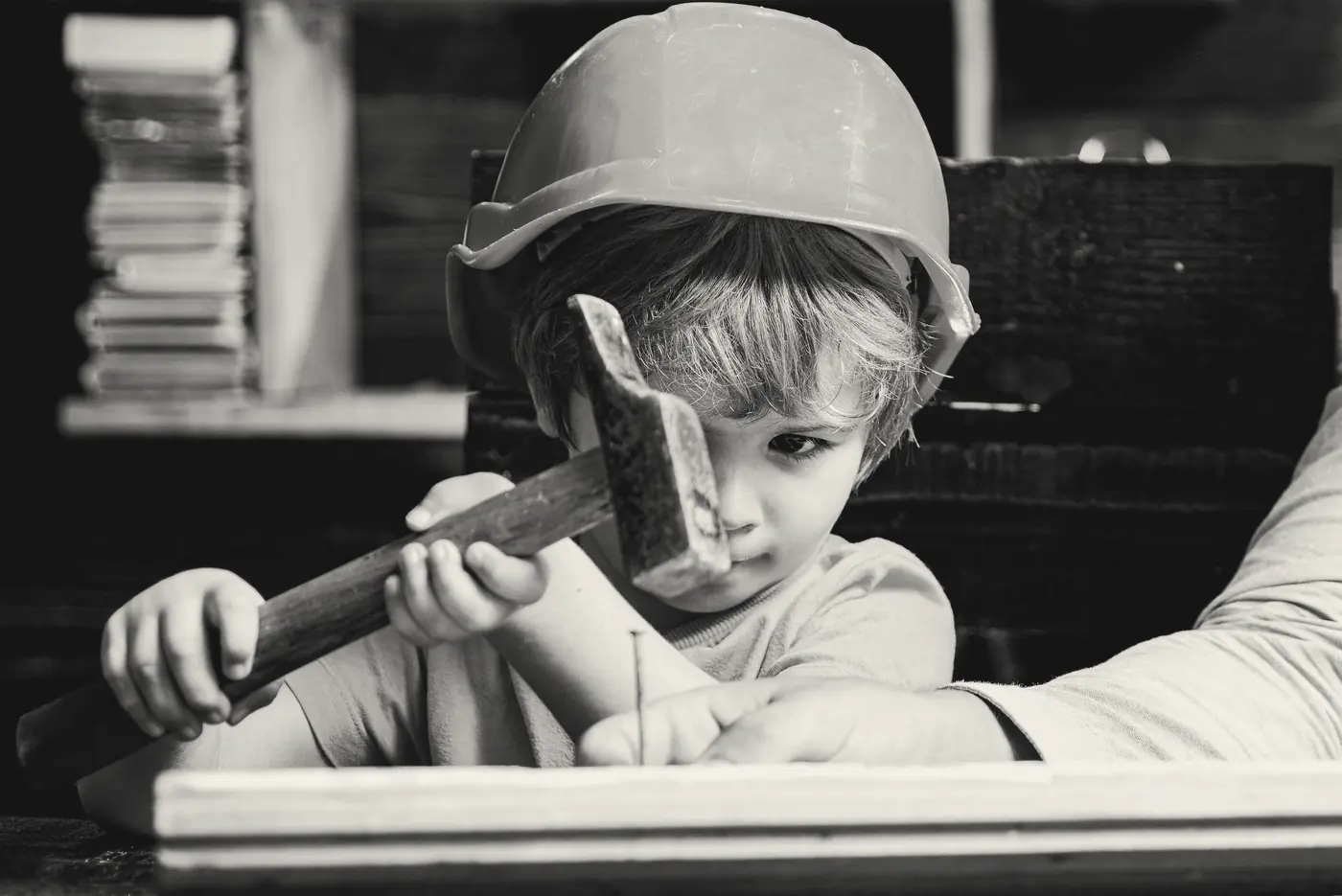 A young child in a hard hat lines up a hammer on a nail while an adult's hand holds the nail steady.
